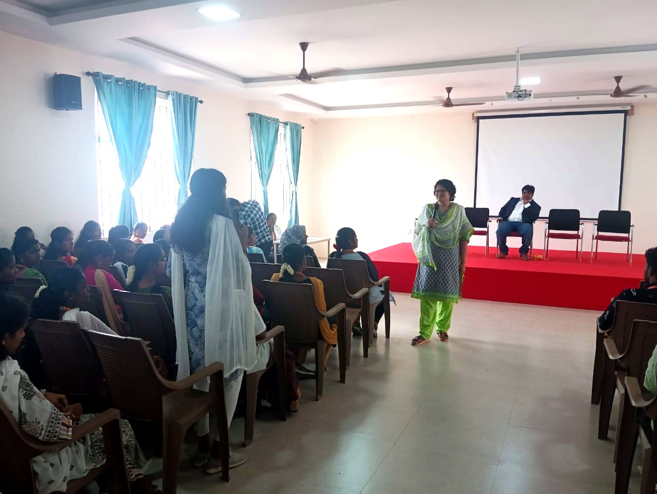 Janani Ramakrishnan speaking to an audience in a bright hall with a red stage